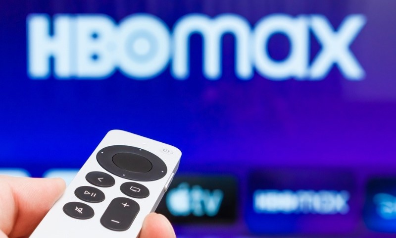 Ruka koja drži beli daljinski upravljač usmeren ka jarko plavom ekranu na kojem se prikazuje zamagljeni logo HBO Max-a